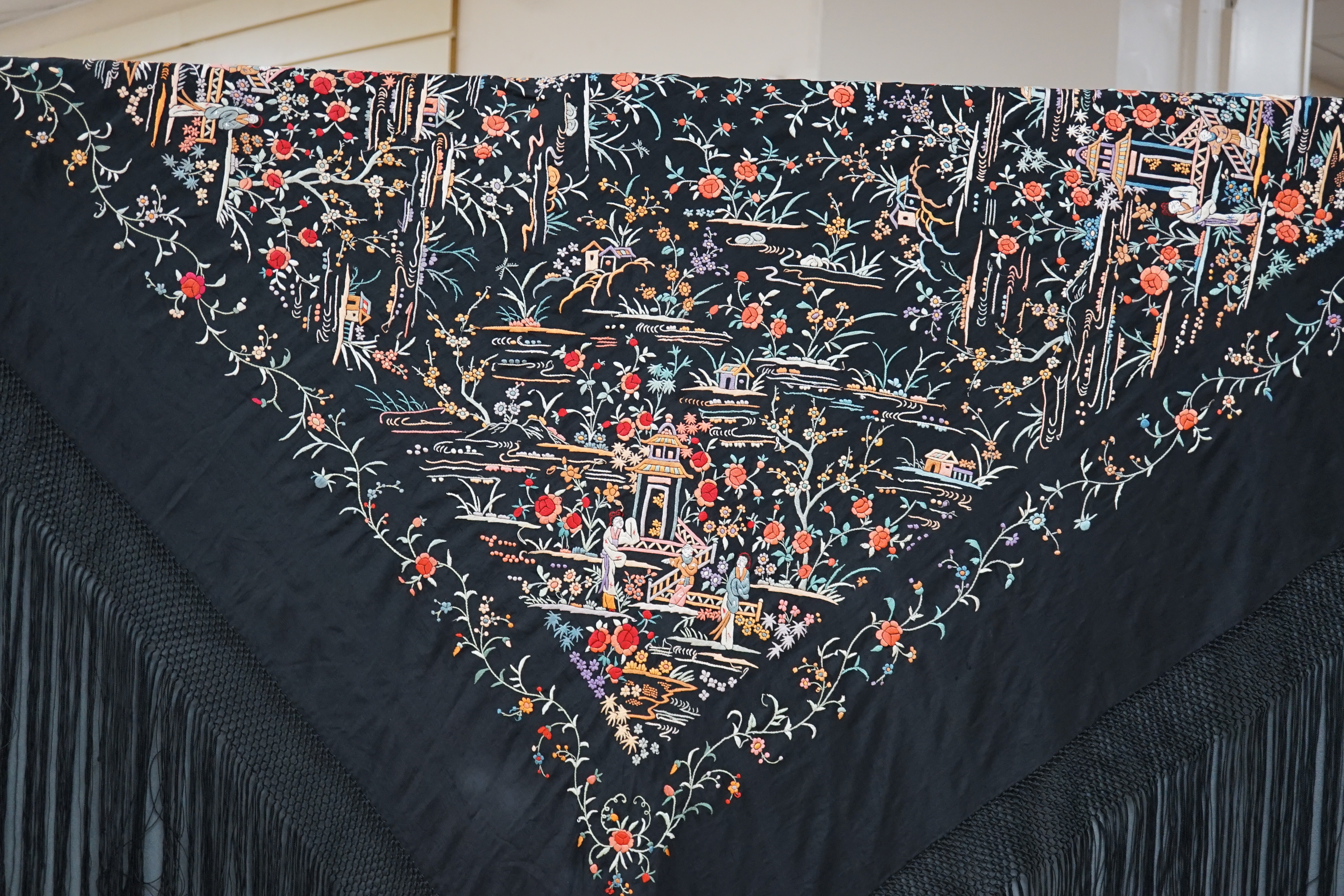 An early 20th century Chinese black silk shawl, with all over coloured floral embroidery, embroidered with multi-coloured silks and a long silk fringe, unusually the four corners are embroidered with a figurative pagoda